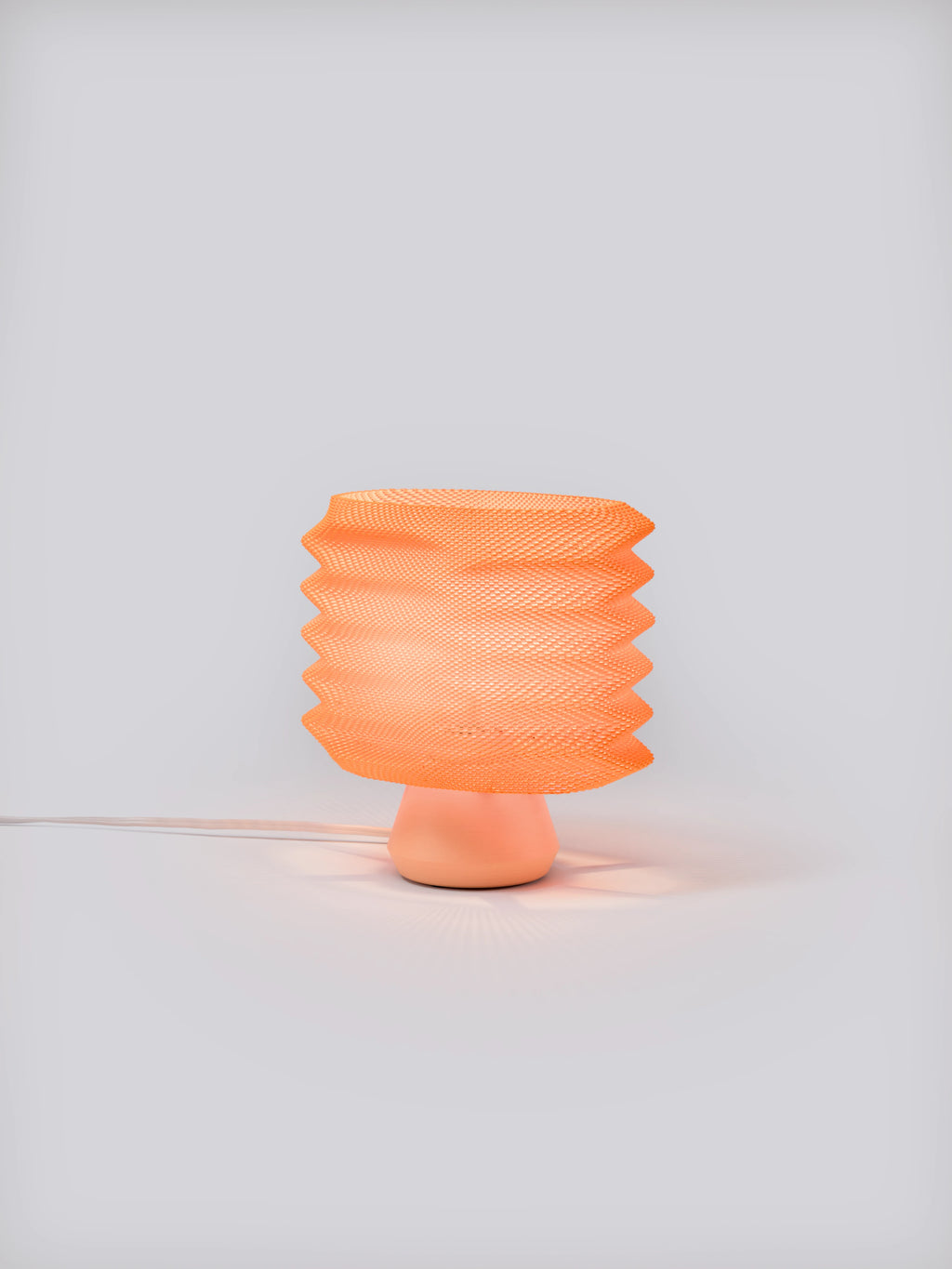 Coral Bellows Weave Lamp illuminated on a white studio background, showing geometric shade and soft glow