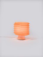 Coral Bellows Weave Lamp illuminated on a white studio background, showing geometric shade and soft glow