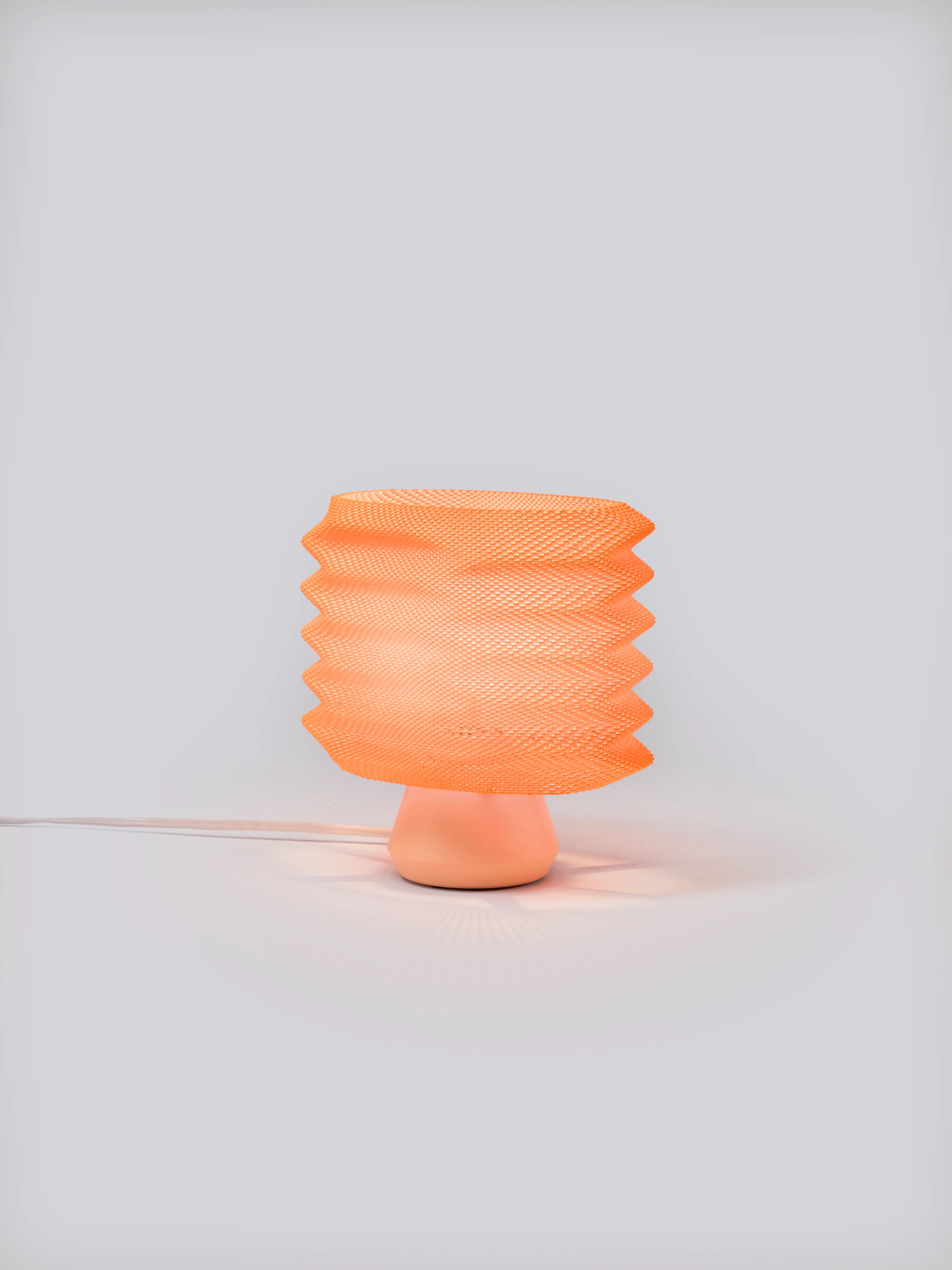 Coral Bellows Weave Lamp illuminated on a white studio background, showing geometric shade and soft glow