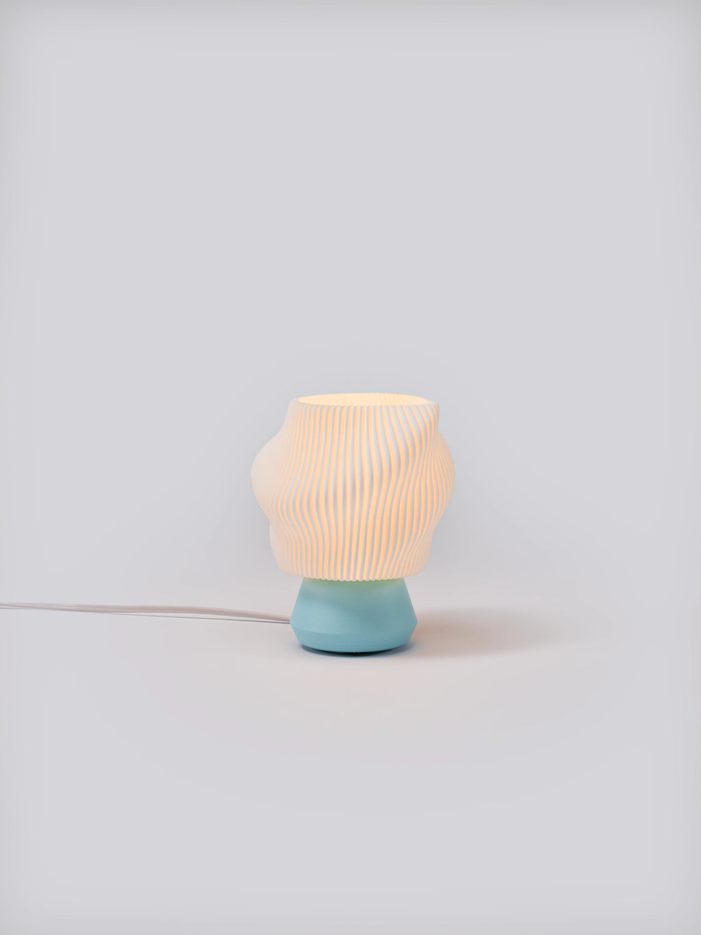 Light blue Dollop Lamp illuminated on a white studio background, showing pleated shade and soft glow