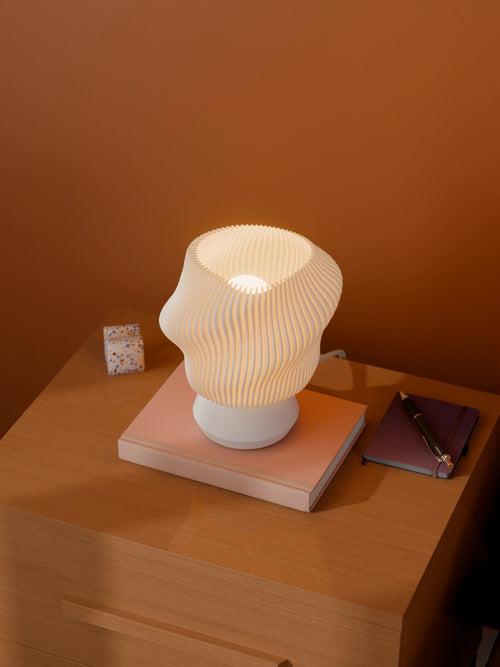 White Dollop Lamp on a wood nightstand, lit, styled with books and a terrazzo block. Viewed from above.