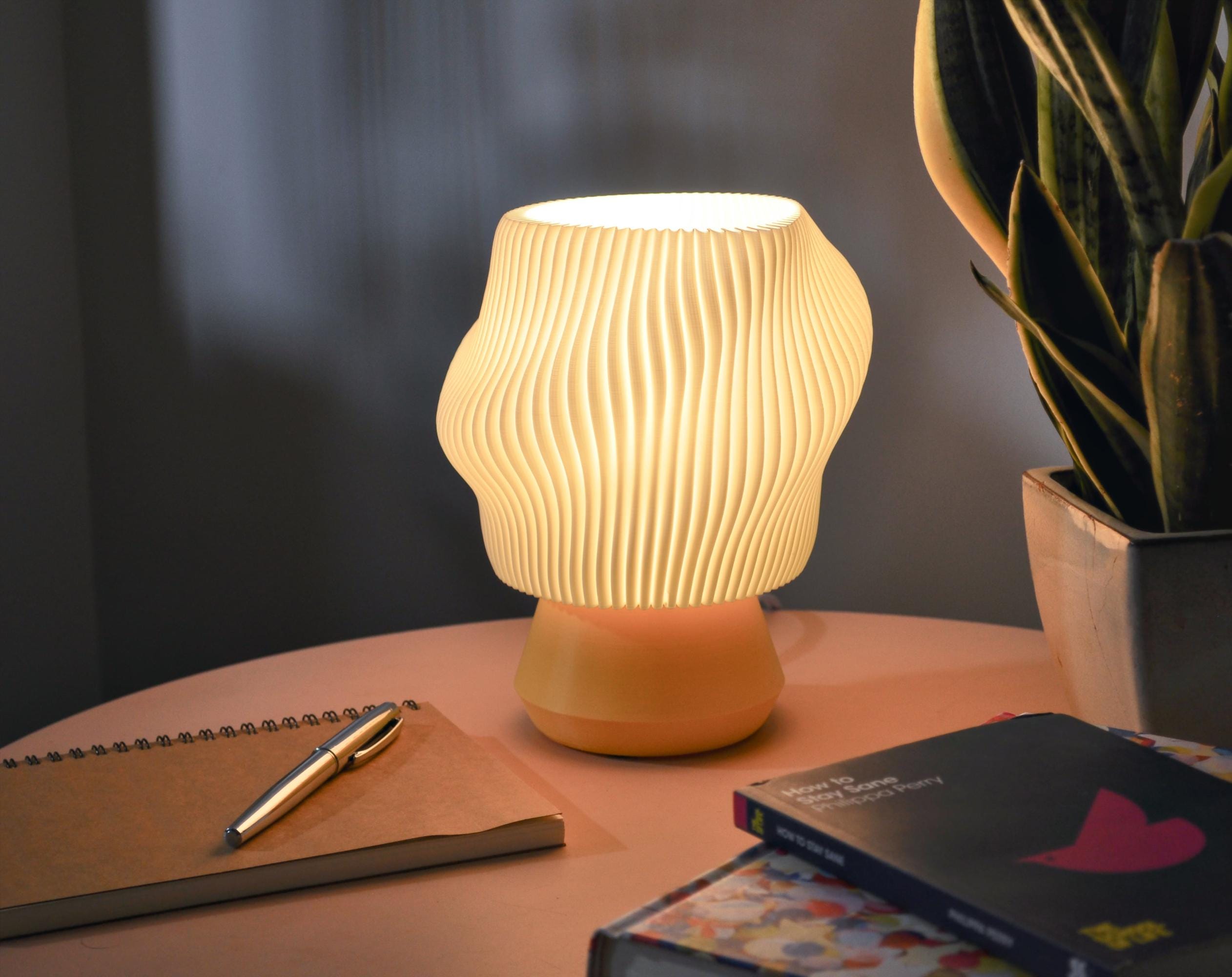 Illuminated wavy pleated white lamp with a yellow base with a plant and books on a table 