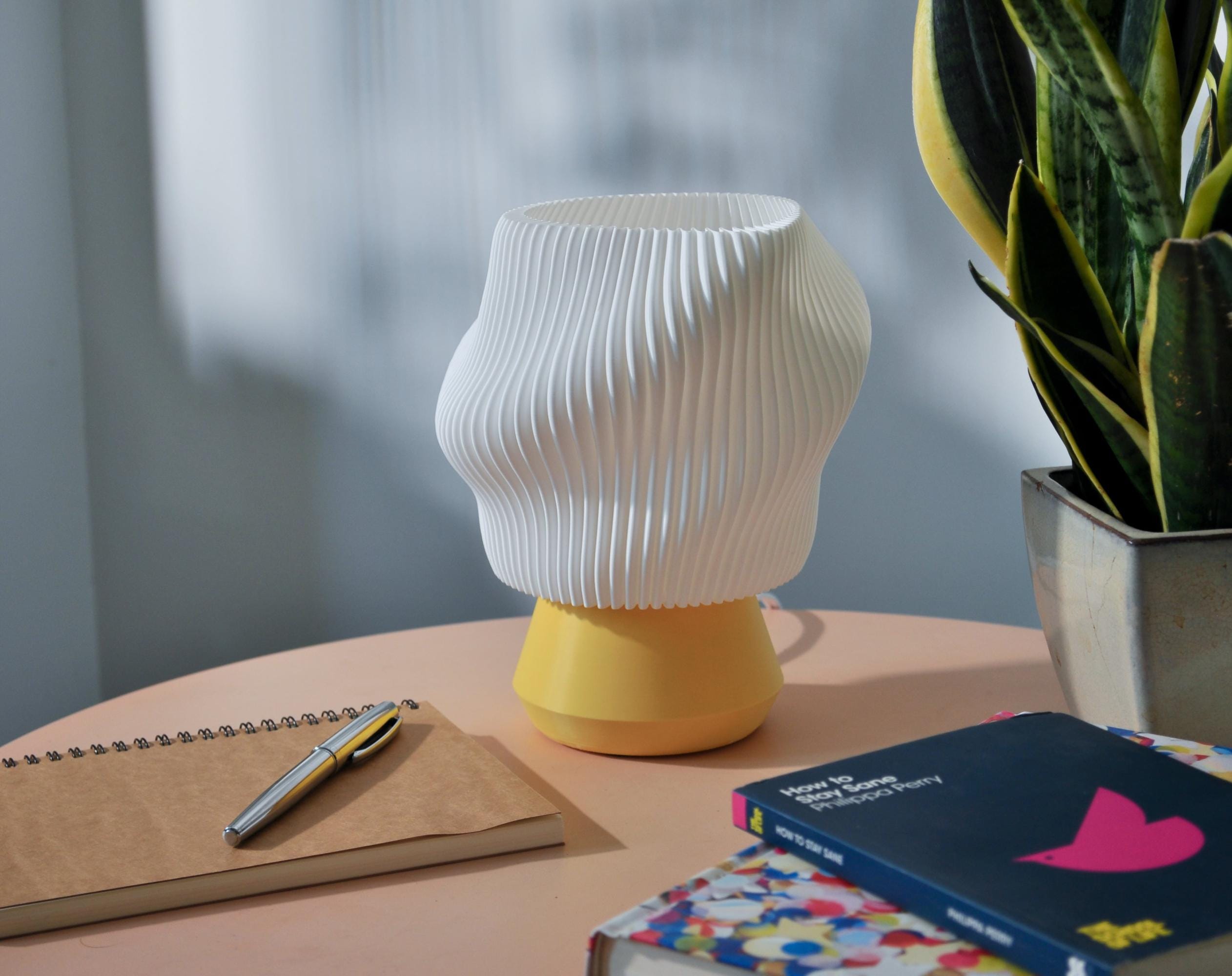 Wavy pleated white lamp with a yellow base with a plant and books on a table