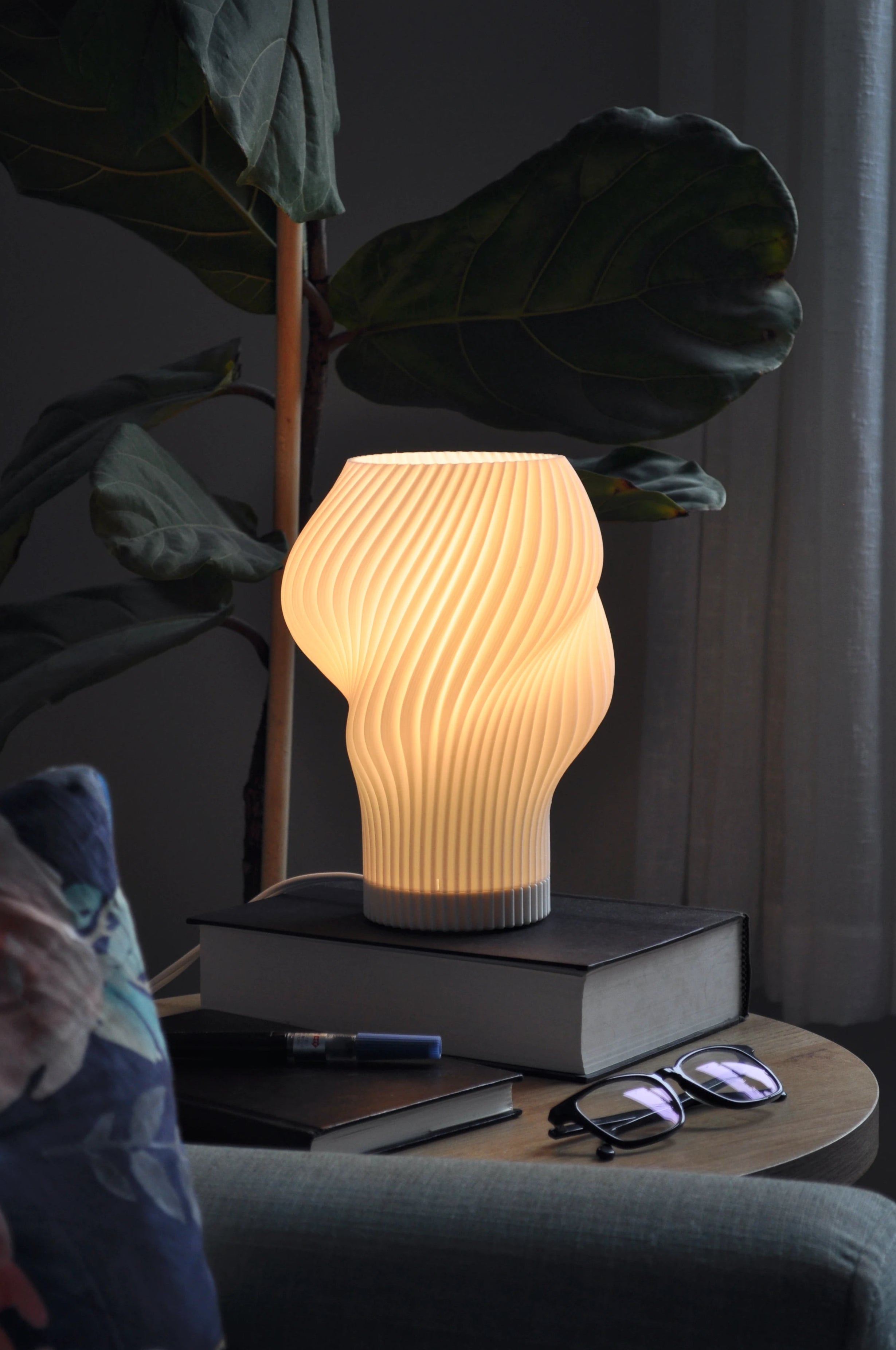Flourish Lamp illuminated on a side table, styled with books and glasses, next to a couch and fiddle leaf fig plant.