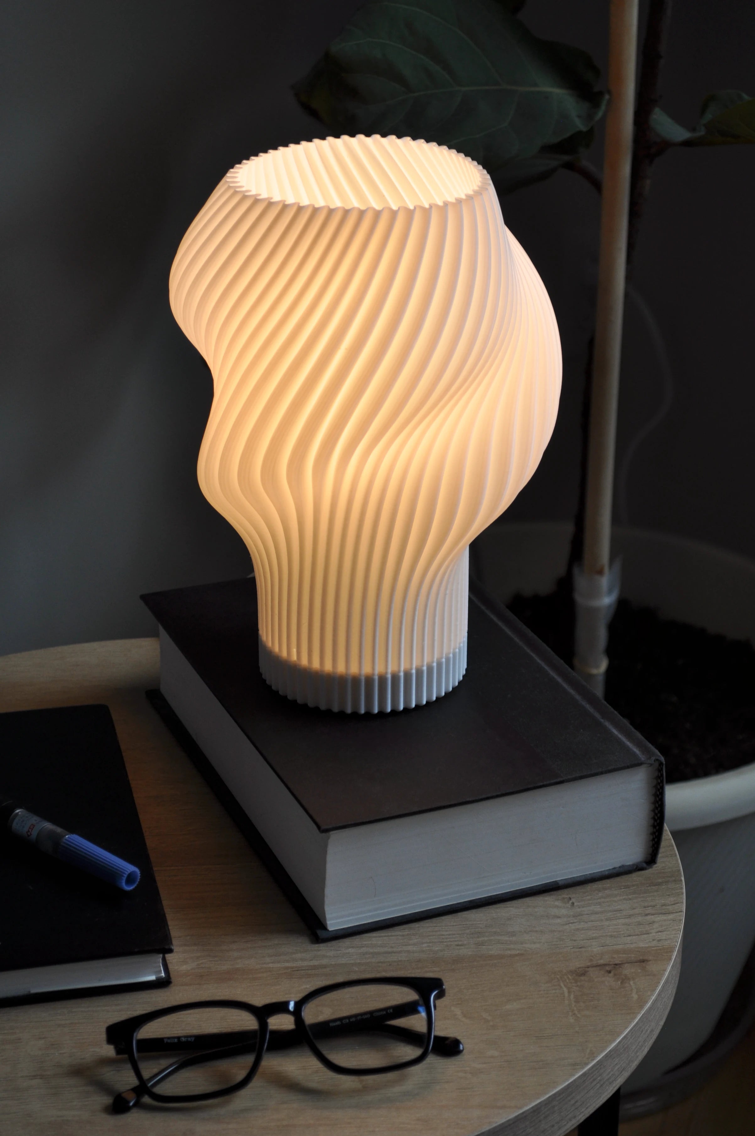 Flourish Lamp illuminated on a side table, styled with books and glasses, next to a couch and fiddle leaf fig plant.
