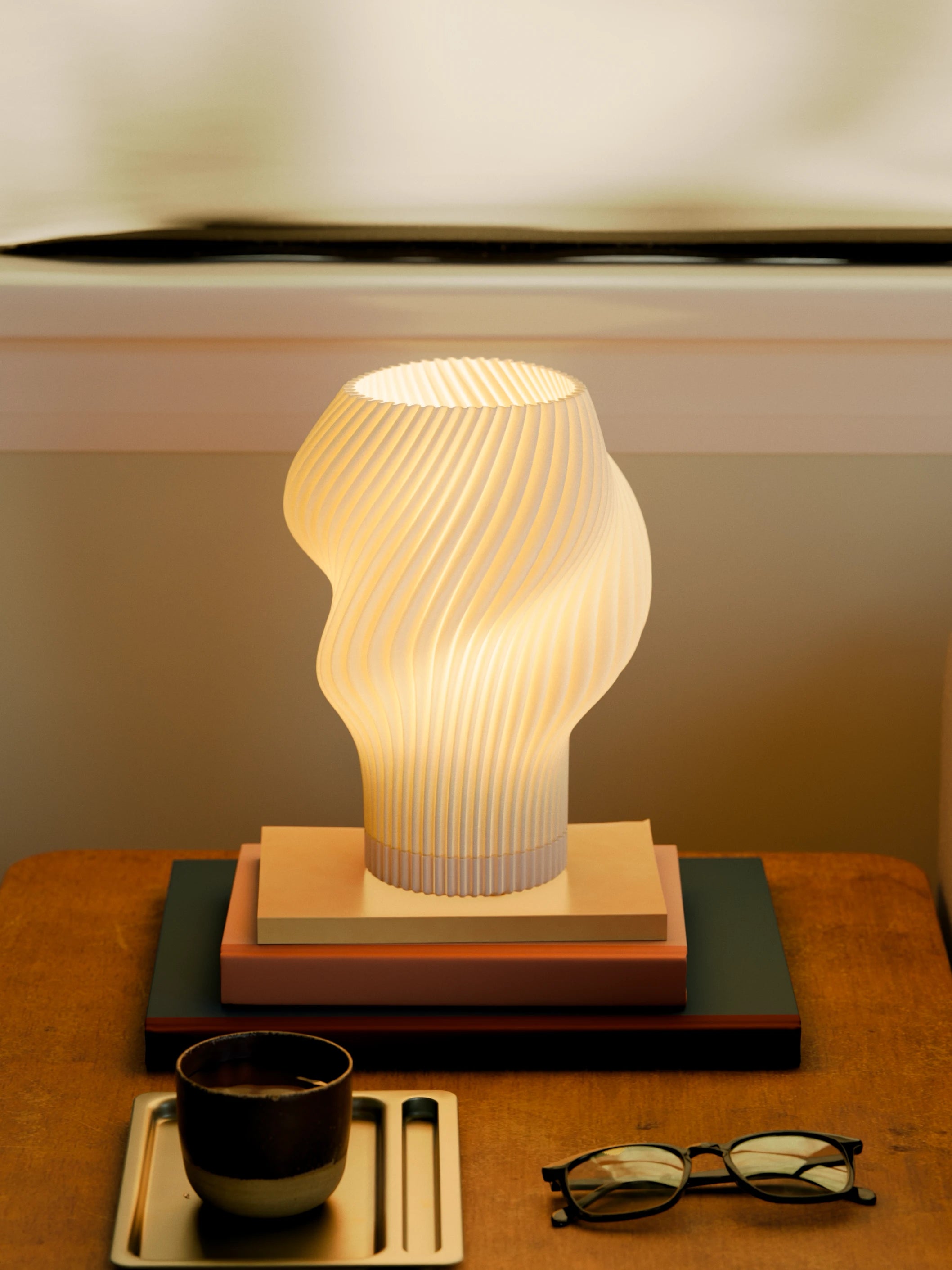 Flourish Lamp illuminated on a side table, styled with stacked books, a coffee cup, and glasses