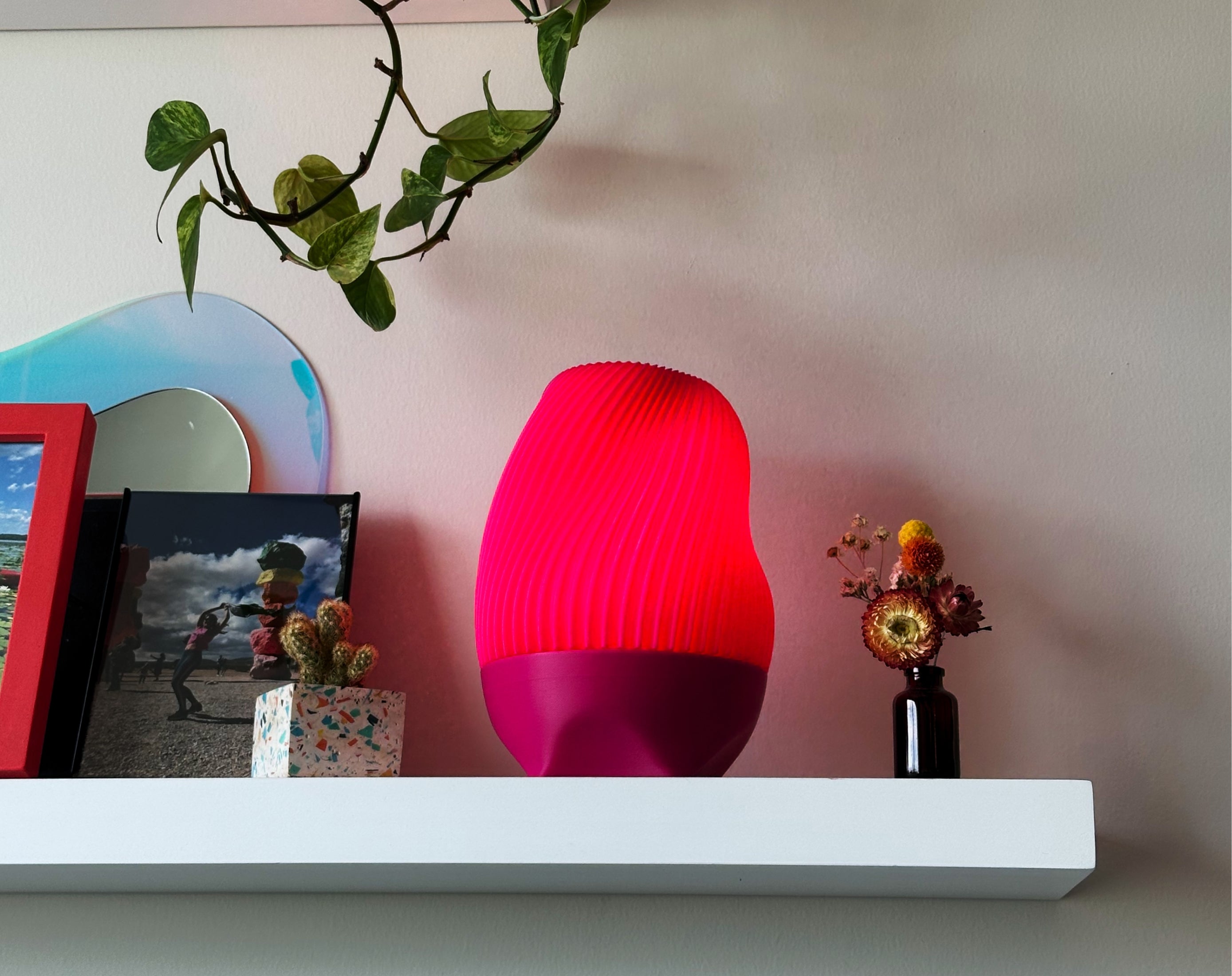 Pink Tama Lamp illuminated on a shelf with photos, a terrazzo vase and plants. 