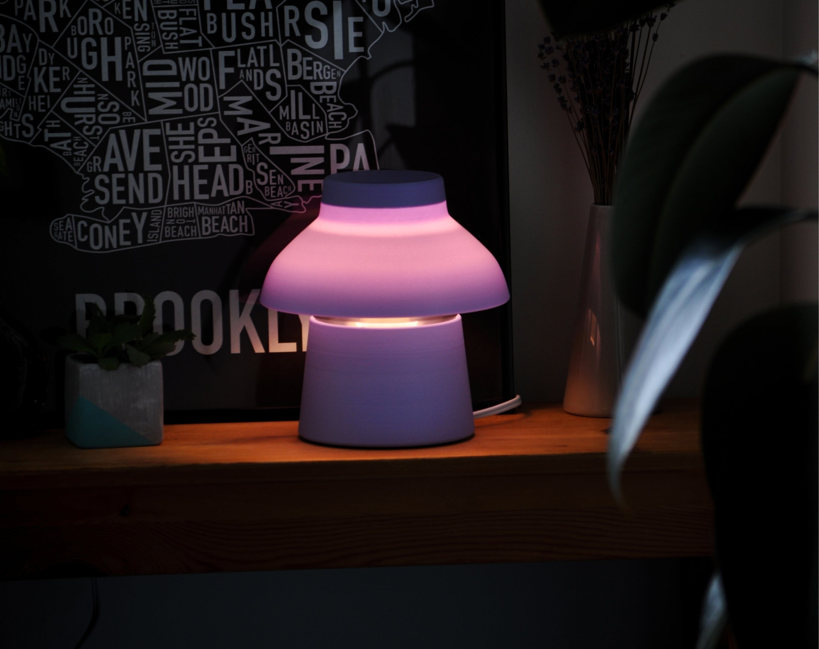 Lavender Zen Lamp illuminated on a table, styled with a poster and plants.