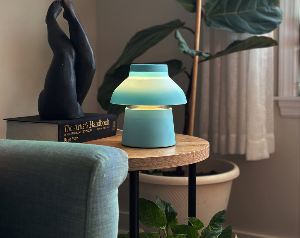 Light Blue Zen Lamp illuminated on a living room side table, styled with a plants, a book, and a bronze sculpture.