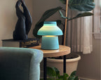 Light Blue Zen Lamp illuminated on a living room side table, styled with a plants, a book, and a bronze sculpture.